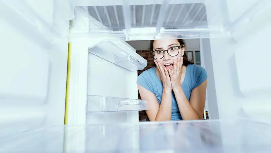 Een vrouw met een blauw shirt en een bril kijkt geschrokken in een lege, witte koelkast met plastic planken.