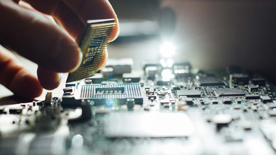 Een hand plaatst een grijze CPU-chip op een groen moederbord met elektronische componenten en reflecterend licht.