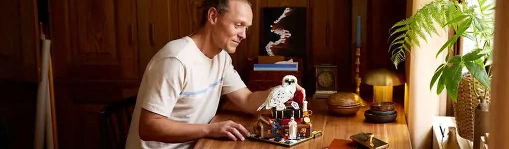 Man zit aan een houten tafel en bouwt rustig een LEGO-model, met planten, lamp en warme houten kastwand op de achtergrond.