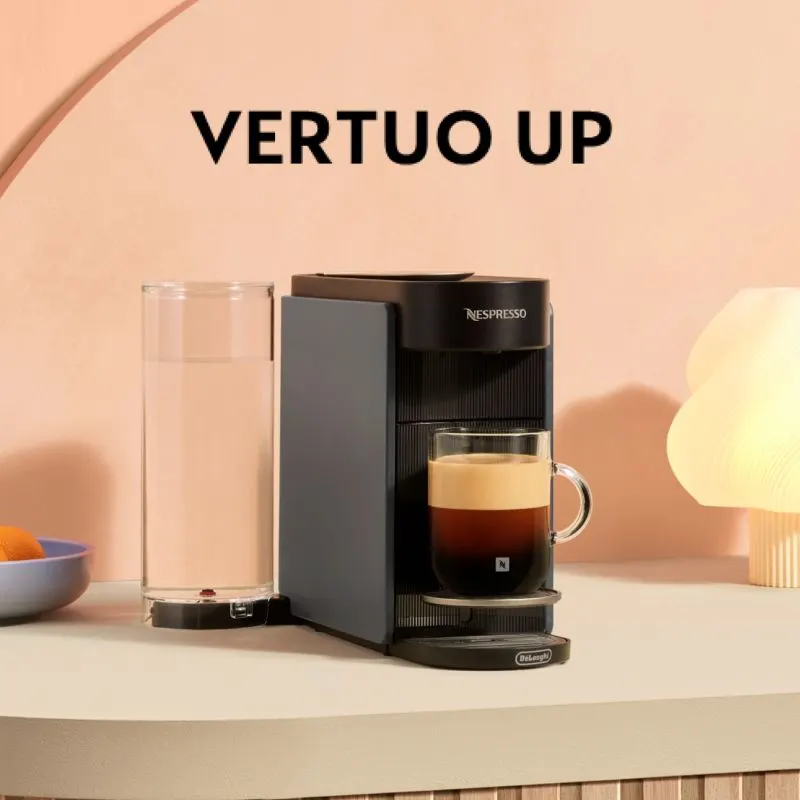 Zwarte Nespresso Vertuo Up koffiemachine met glazen kop koffie en waterreservoir op een beige tafel tegen perzikkleurige achtergrond.