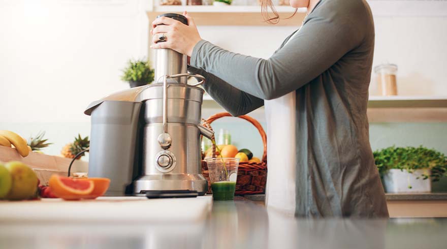 Een vrouw perst sap met een zilveren slowjuicer in de keuken, met fruit en een groene drank in de buurt.