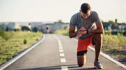 Een man in een grijs shirt en oranje korte broek kijkt op een pad naar zijn smartwatch. Op de achtergrond groen en gebouwen.