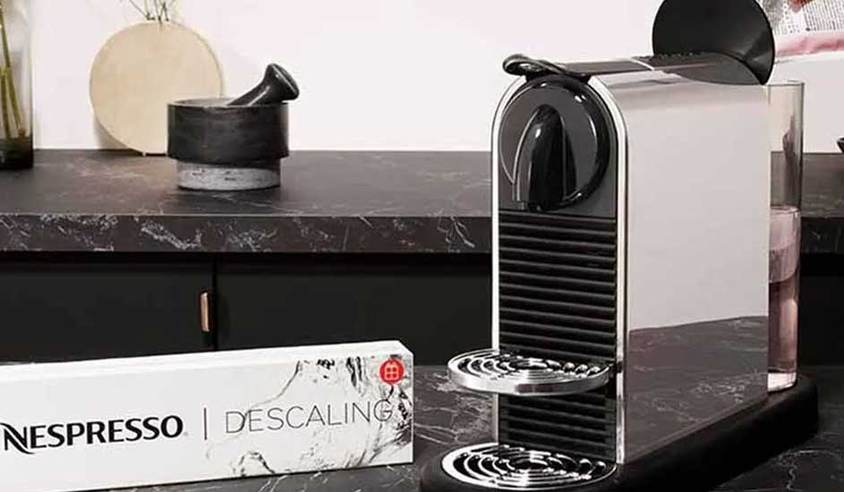 Op een zwart marmeren aanrecht staat een zilveren Nespresso-apparaat naast een doosje ontkalkingsmiddel in een moderne keuken.