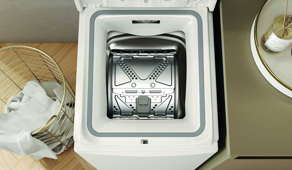 Een bovenlader wasmachine zonder zichtbaar merk staat open, naast een wasmand met wasgoed en een kastje met een kaars erop.