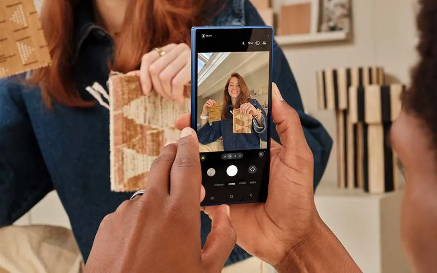 A smartphone camera photographs someone holding fabric or wallpaper samples, with the shot clearly displayed on the screen.
