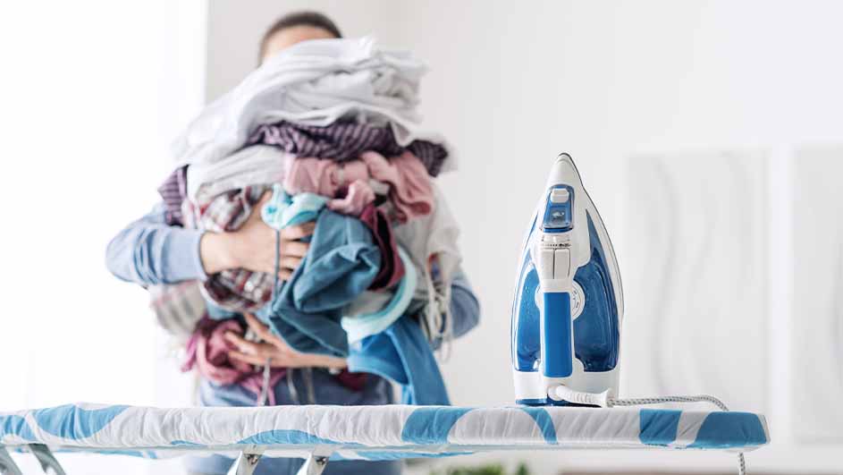 Een persoon draagt een stapel wasgoed achter een blauw-wit gestreekt strijkplank met een blauw-wit strijkijzer erop.