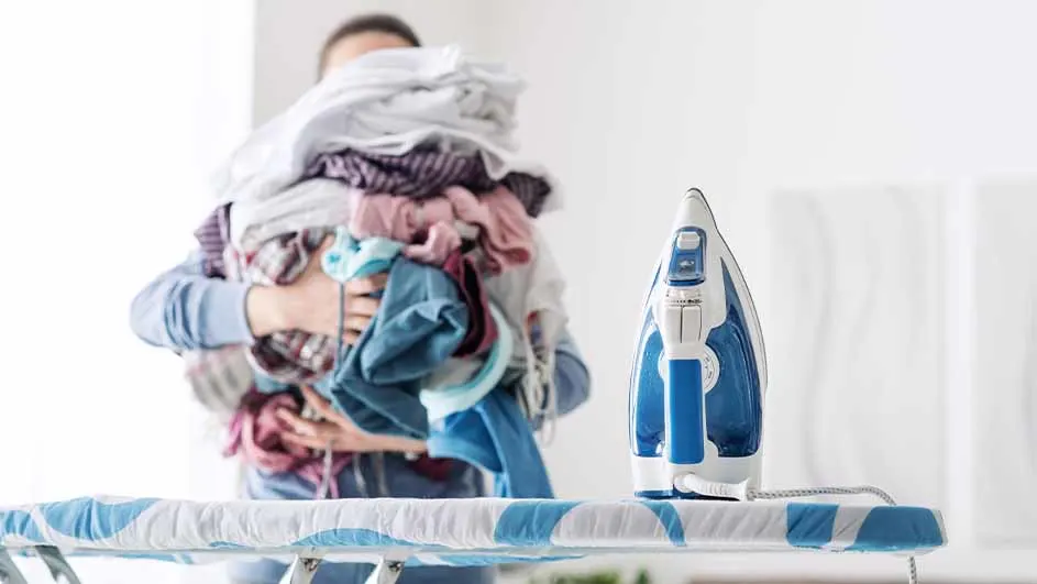 Een persoon draagt een stapel wasgoed achter een blauw-wit gestreekt strijkplank met een blauw-wit strijkijzer erop.