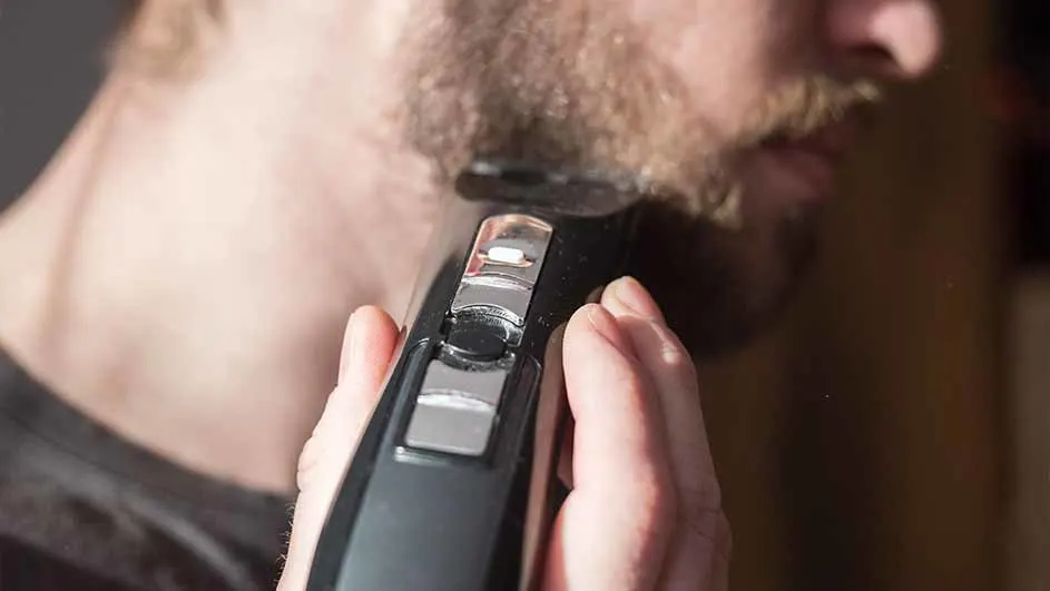 Een man trimt zijn baard met een zwarte, elektrische trimmer met zilverkleurige knoppen. De achtergrond is wazig.