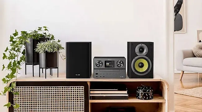On a wooden cabinet stands a black Philips stereo set with two speakers, next to green plants and books in a modern interior.