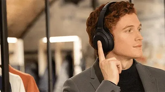 A person is wearing black headphones and a gray blazer. Their finger is touching the headphone. The background is blurred.