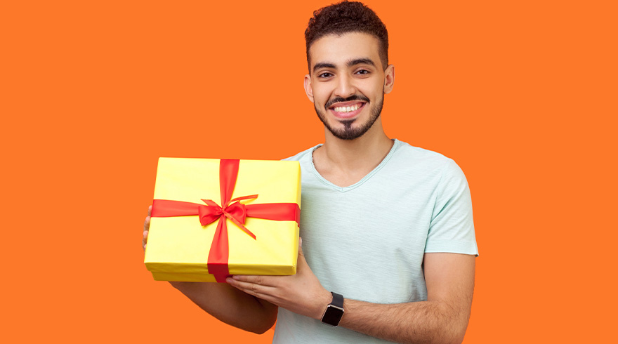 Een man glimlacht en houdt een geel cadeau vast met een rode strik. De achtergrond is oranje. Hij draagt een lichtblauw t-shirt.