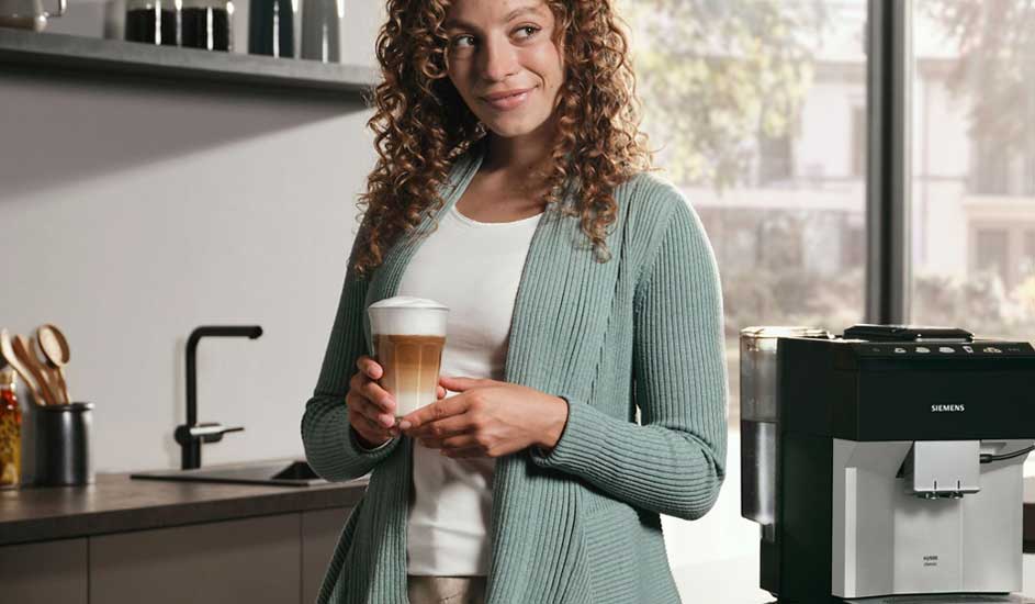 Een vrouw staat in een moderne keuken en houdt een glas latte macchiato vast, naast een volautomatische koffiemachine van Siemens.