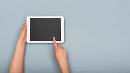 Twee handen houden een witte tablet met een zwart scherm vast tegen een blauwe achtergrond. Een vinger raakt het scherm aan.