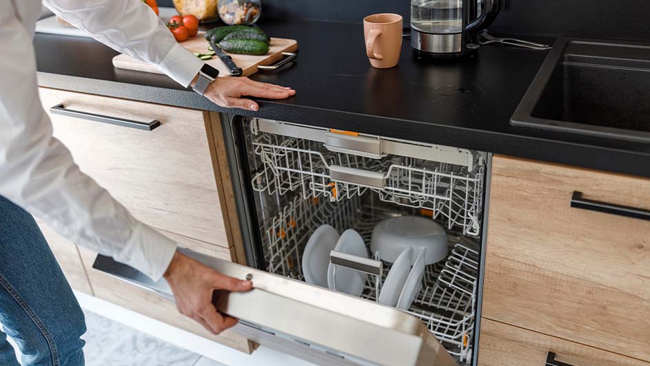 Een persoon opent een vaatwasser in een keuken met houten kasten, een zwarte aanrecht en keukengerei.
