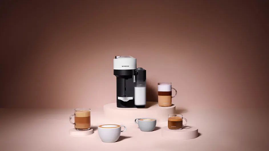 Een koffiezetapparaat met melkopschuimer staat met diverse kopjes koffie op een podium. De achtergrond is roze.