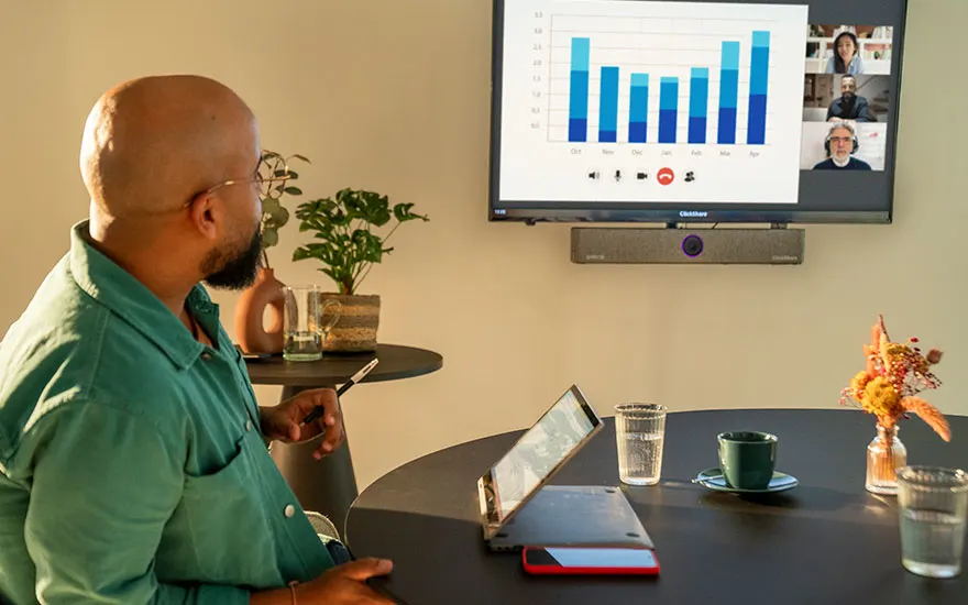 Een collega in groen shirt tijdens videovergadering met laptop, bekijkt blauwe staafgrafiek op wandscherm terwijl andere deelnemers meekijken.