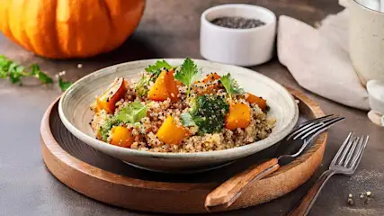 Een kom quinoa-salade met pompoen en peterselie op een houten bord, met een pompoen en bestek op de achtergrond.