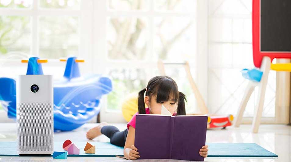 Een kind ligt op een mat in een lichte kamer en leest een paars boek, naast een witte luchtreiniger en wat kleurrijk speelgoed.