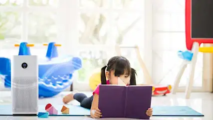 Een kind ligt op een mat in een lichte kamer en leest een paars boek, naast een witte luchtreiniger en wat kleurrijk speelgoed.