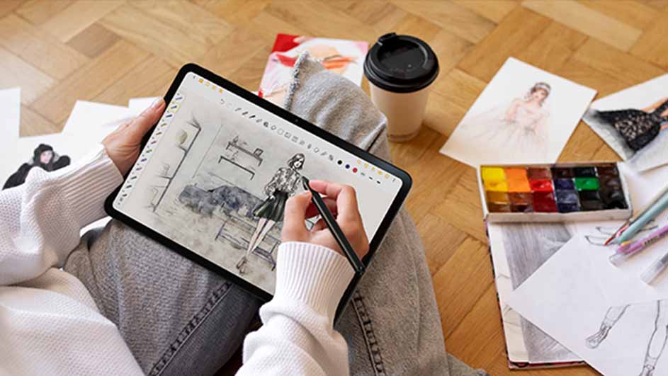 Een persoon tekent een mode-illustratie op een tablet met een pen, omringd door schetsen, waterverf en een beker op een houten vloer.