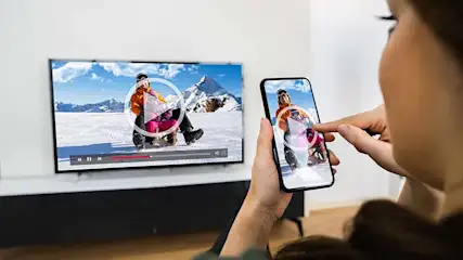 Vrouw kijkt video op telefoon; tv toont dezelfde video van iemand op een slee in een besneeuwd berglandschap.