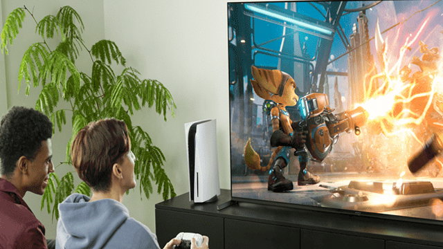 Twee mensen spelen een videospel op een tv naast een groene plant en een witte spelcomputer.