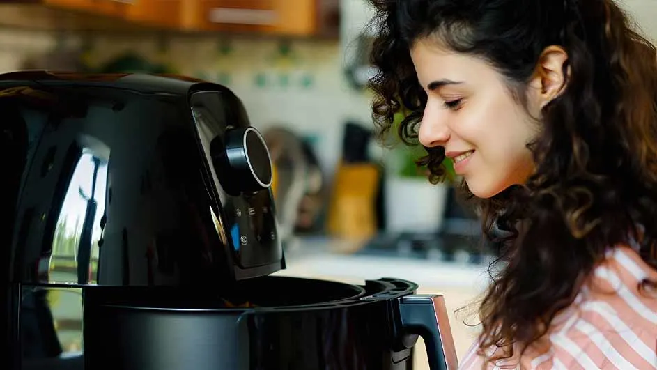 Een vrouw kijkt naar een zwarte airfryer in een keuken, met houten kasten op de achtergrond.