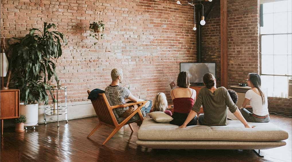 Groep mensen kijkt tv in een kamer met een bakstenen muur, houten vloer en grote planten.