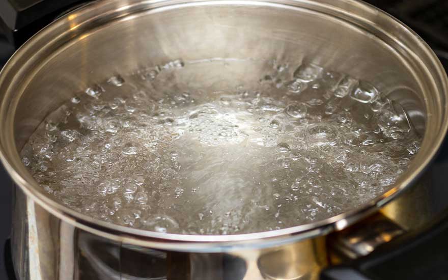 A clear glass kettle with a black handle and base sits on a white table, filled with boiling water. A grey cabinet is in the background.