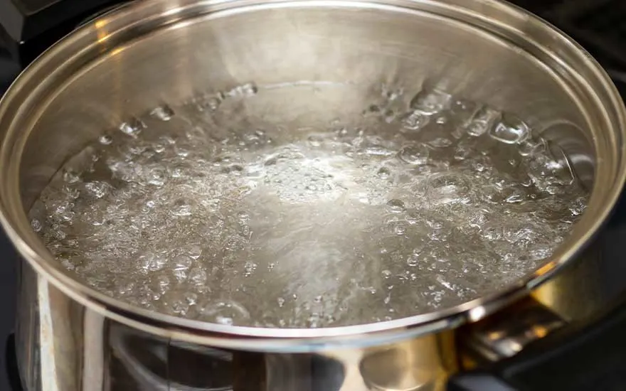 A clear glass kettle with a black handle and base sits on a white table, filled with boiling water. A grey cabinet is in the background.