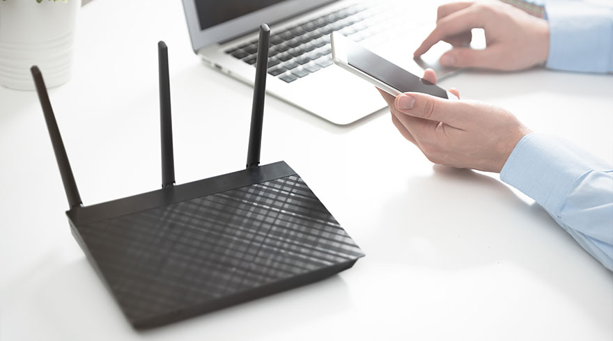 Zwarte router met drie antennes op een witte tafel, met een laptop en handen met een smartphone.
