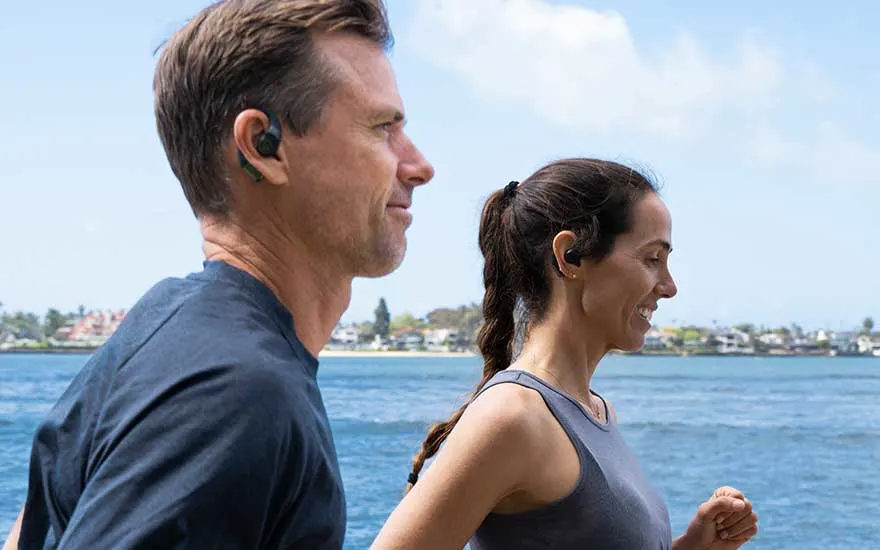 Two people with wireless earbuds jog along blue water with a coastline in the background under a clear sky.