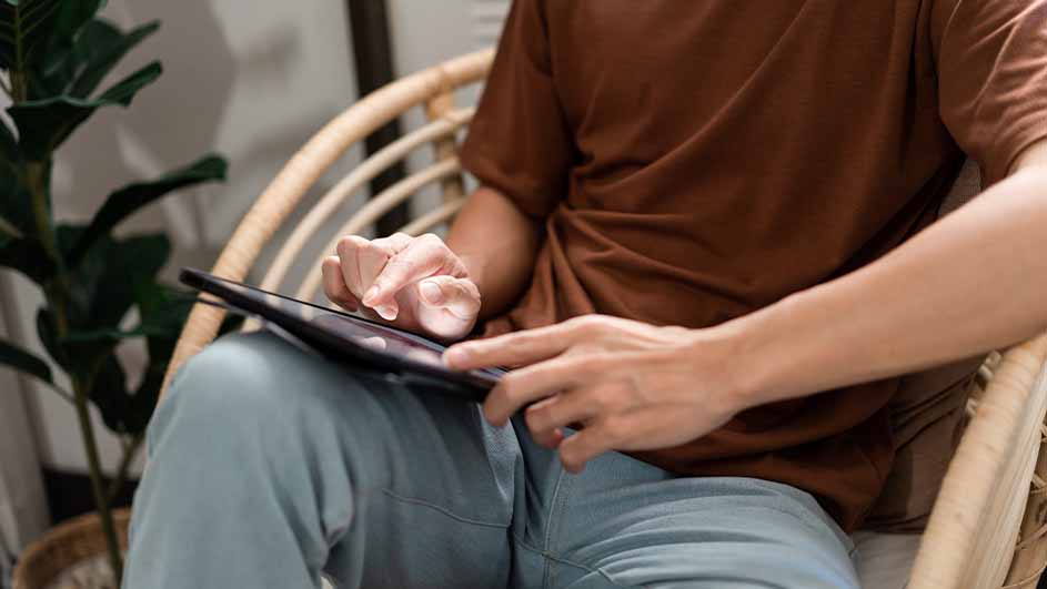 Een persoon in bruin shirt en blauwe broek zit in een rieten stoel en gebruikt een tablet. Op de achtergrond staat een plant.