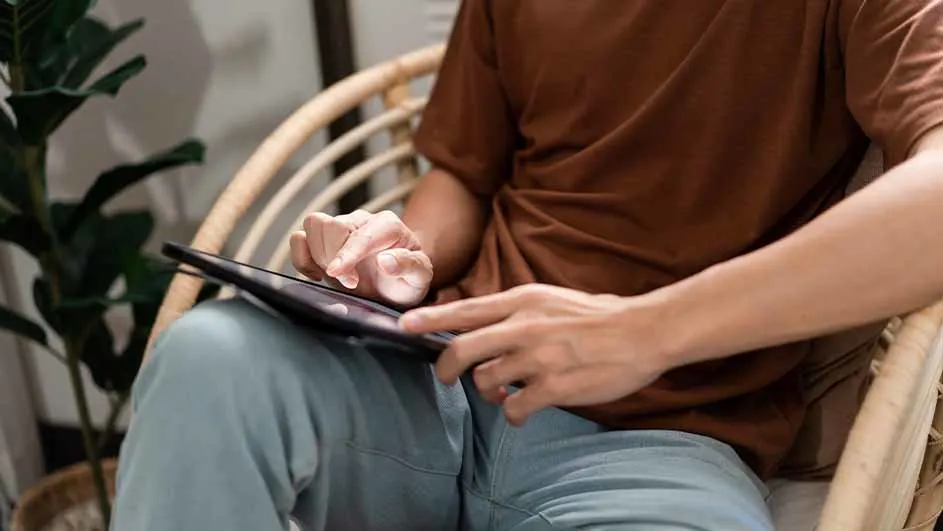 Een persoon in bruin shirt en blauwe broek zit in een rieten stoel en gebruikt een tablet. Op de achtergrond staat een plant.