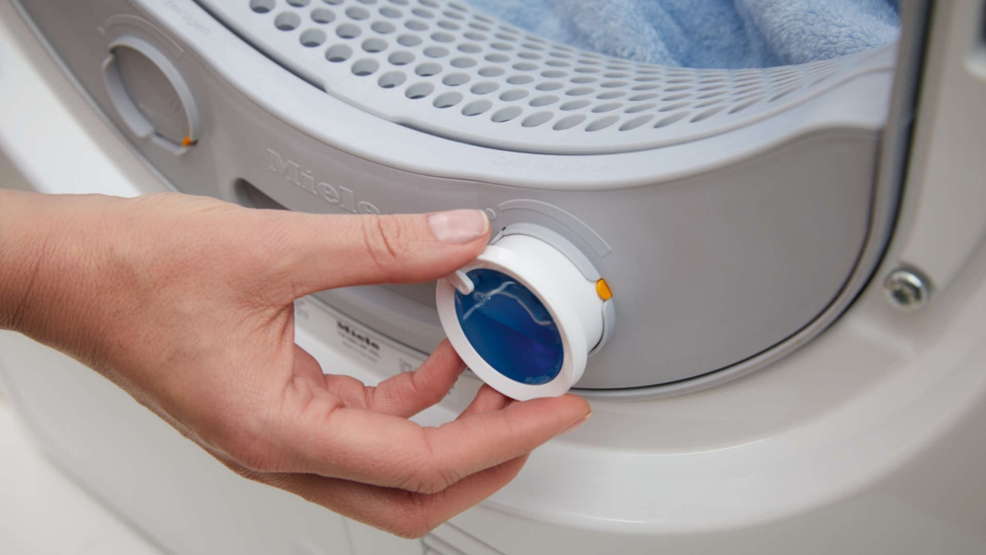 Een hand draait aan een blauw-witte knop van een witte Miele droger, met blauw wasgoed zichtbaar binnenin.