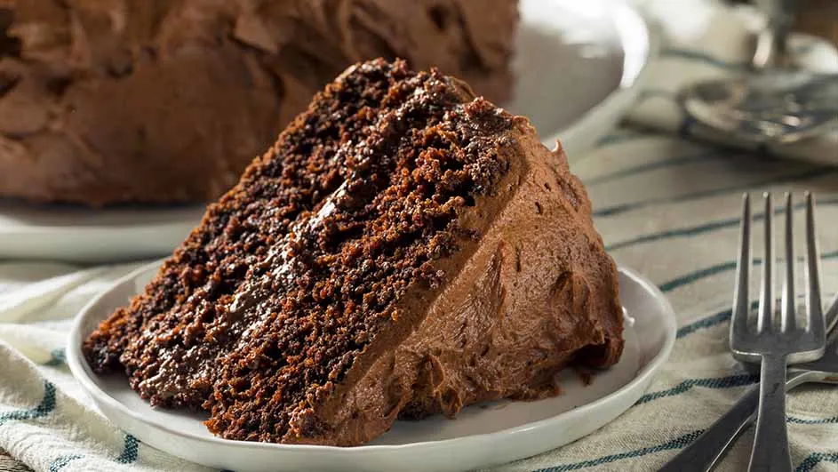 Een stuk chocoladetaart op een wit bord met een vork ernaast, op een gestreepte doek. Op de achtergrond een grotere taart.