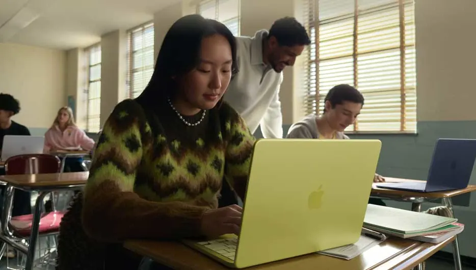 Leerlinge typt geconcentreerd op een gele laptop in het klaslokaal, terwijl de leraar naast haar buigt en klasgenoten achteraan meewerken.