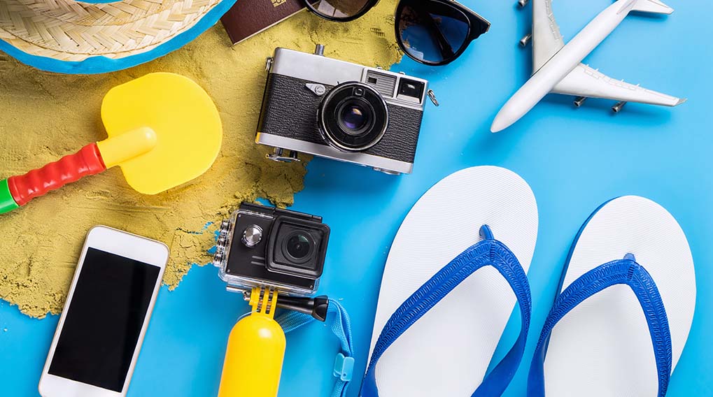 Vakantie-items op blauwe achtergrond: camera's, telefoon, paspoort, zonnebril, flip-flops, vliegtuig, hoed, zandspeelgoed en zand.