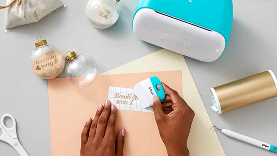 Ambachtelijke tafel met blauwe Cricut machine, gouden vinylrol, doorzichtige kerstballen en handen die een sticker aanbrengen.