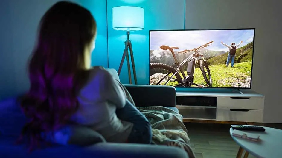 Woman on sofa watches a large TV showing nature and a mountain bike; blue-purple lamp glow. No brand visible.