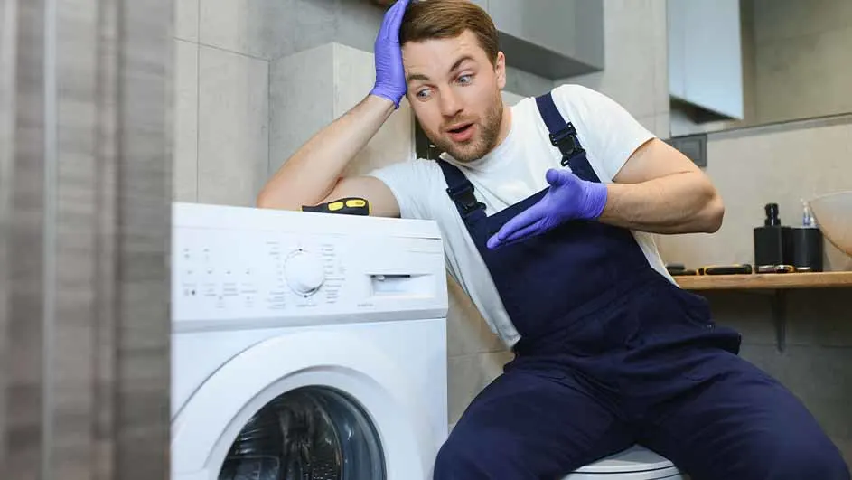 In a bathroom setting, a repairman in blue overalls and gloves leans against a white washing machine with a surprised expression.