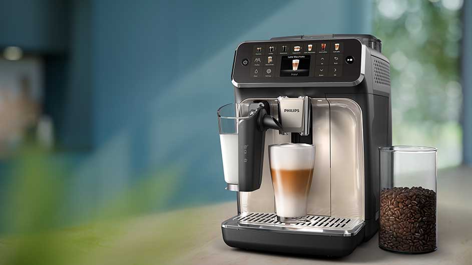Close-up van een Philips volautomatische espressomachine die melk opschuimt en een latte macchiato bereidt, met een glazen pot koffiebonen ernaast.