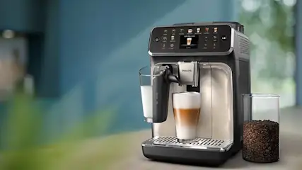 Close-up van een Philips volautomatische espressomachine die melk opschuimt en een latte macchiato bereidt, met een glazen pot koffiebonen ernaast.