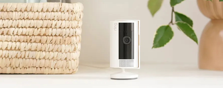 Een witte Ring camera staat op een witte tafel, naast een rieten mand en een vaas met groene bladeren.