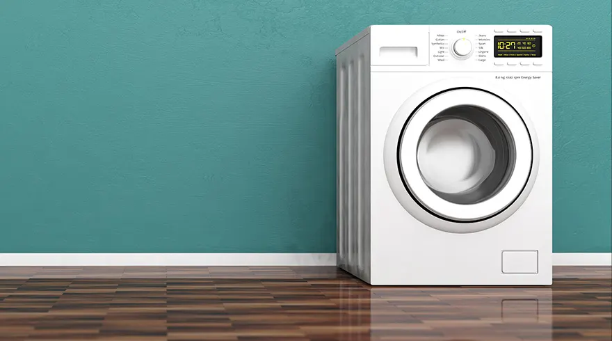 Een witte wasmachine staat tegen een blauwe muur op een houten vloer. Het display toont 10:27.