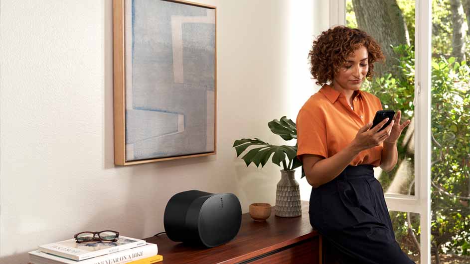 Vrouw staat bij het raam met een telefoon. Naast haar staat een kast met een speaker, plant, boeken en een bril.