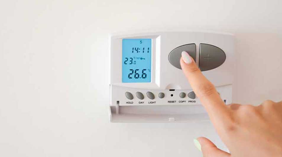 A finger presses a gray button on a white thermostat mounted on a white wall. The display reads 14:11 and 26.6 degrees.