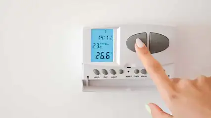 A finger presses a gray button on a white thermostat mounted on a white wall. The display reads 14:11 and 26.6 degrees.