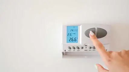 Een witte thermostaat aan een witte muur toont 14:11 en 26,6 graden. Een vinger drukt op een grijze knop.