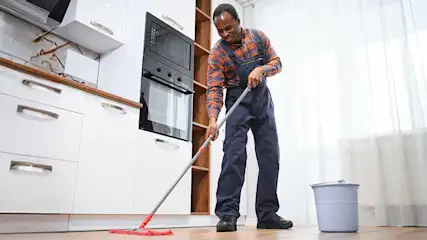 Een man dweilt een keuken met witte kasten, een oven, een houten aanrecht en een grijze emmer.